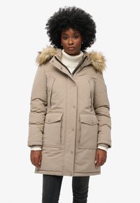 Parka beige con cappuccio foderato in pelliccia, dotato di chiusura a zip e a scatto, due tasche frontali e polsini a coste. Texture liscia con isolamento imbottito.