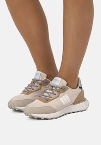 Beige Sportschuhe mit Obermaterial aus Wildleder und Mesh, gemusterten Schnürsenkeln, strukturiertem Mittelsohlenbereich und ausgeprägter Gummiaußensohle. Markenlogo seitlich sichtbar.