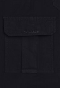 Musta kangas, jossa on suorakaiteen muotoinen tasku ja läppä. Sana "FREDDY" on brodattu taskun alle. Yksinkertainen muotoilu, sileä rakenne.