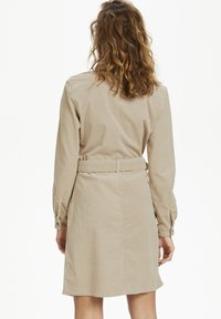 Abito camicia in corduroy color moka con maniche lunghe, vita a corsetto e taglio dritto. Presenta un tessuto testurizzato e dettagli con bottoni ai polsini.