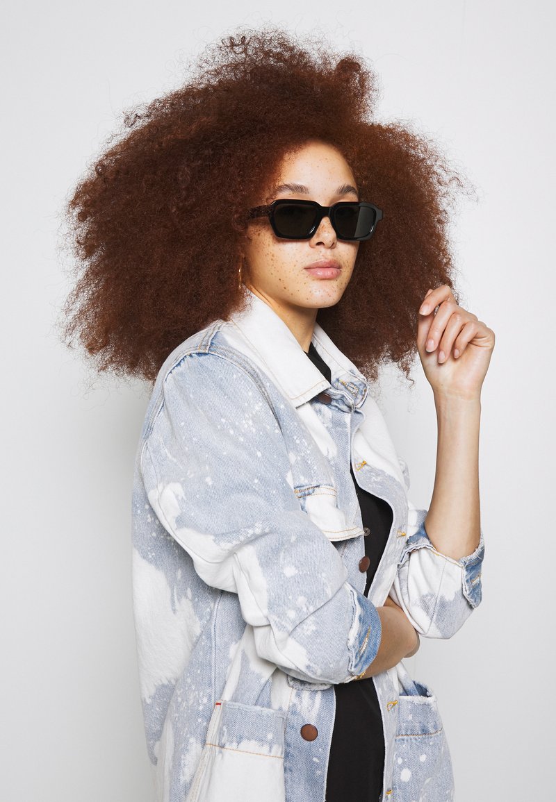 Lunettes de soleil noires rectangulaires portées avec une veste en denim bleu clair et blanche éclaboussée. Des cheveux roux bouclés et volumineux entourent le visage.