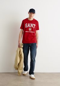 Tricou roșu GANT cu logo alb, blugi albastri, jachetă crem în mână și șapcă neagră. Tenisi albi completează ținuta. Fundal simplu.