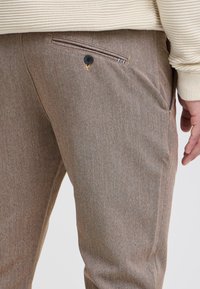 Hnědé, texturované kalhoty s boční kapsou se černým knoflíkem a jemným vzorem. Materiál je pravděpodobně směs vhodná pro běžné nošení.