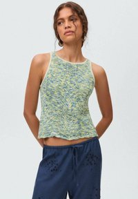 Knitted tank top in yellow and blue with a scalloped hem, sleeveless design, and a textured pattern. Paired with navy blue pants.