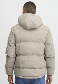 Beige puffer jacket with a hood, featuring a quilted design and elastic cuffs. Smooth exterior and padded for insulation.