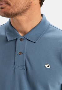 Blå pikétröja i strukturerad tyg, med klassisk krage, två bruna knappar och en liten logotyp på bröstet.