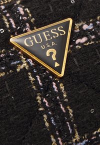 Tissu en tweed noir avec des fils multicolores. Logo triangulaire doré et noir proéminent avec "GUESS" et un point d'interrogation.