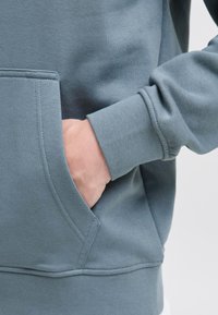 Hætte sweatshirt i dæmpet teal stof med en frontlomme og ribbede manchetter. Har en glat tekstur og en ren syning.