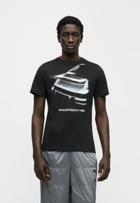 Μαύρο βαμβακερό μπλουζάκι με λευκό γραφικό της Porsche στο μπροστινό μέρος, με λαιμόκοψη και κοντά μανίκια. Φορεμένο με γκρι σορτς.