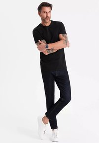 Černé tričko s krátkým rukávem, tmavě modré džíny a bílé tenisky. Obsahuje viditelné tetování a stříbrné hodinky. Uvolněný, přiléhavý design.