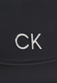 Črna usnjena površina z izstopajočim srebrnim logotipom "CK". Gladka tekstura s subtilno zrnatostjo in natančnim šivanjem.