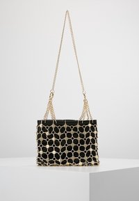 Bolso negro con cadena dorada y revestimiento de malla metálica dorada, exhibido en un pedestal blanco contra un fondo gris.