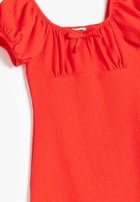Haut texturé rouge avec des manches bouffantes courtes et un col en U, présentant des détails froncés aux épaules et un accent à nouer.