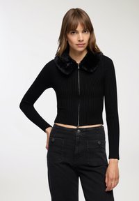 Kobieta w czarnym, prążkowanym swetrze z długim rękawem, zapinanym na zamek, z krótkim krojem i sztucznym futrem przy kołnierzu, zestawionym z czarnymi spodniami o wysokim stanie z przednimi kieszeniami.