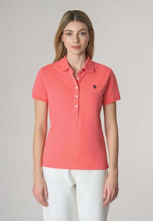 Polo shirt corallo con maniche corte e colletto, con un logo in blu navy. Bottoni lungo la parte anteriore, abbinato a pantaloni bianchi.