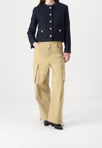 Woman wearing navy blue buttoned jacket and beige wide-leg cargo pants with black heeled boots, standing with one hand in pocket.