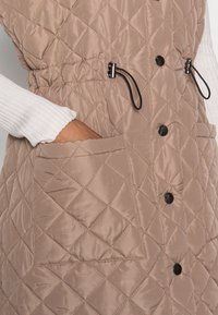 Gilet matelassé beige avec boutons noirs, cordons de serrage ajustables et poches avant. Texture lisse et motif en losange sur toute la surface.