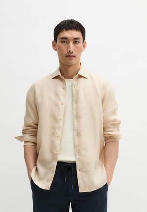 Homme aux cheveux noirs courts portant une chemise beige ouverte sur un top blanc texturé et un pantalon foncé à cordon, debout devant un fond uni.