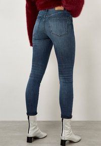 Persona con jeans ajustados azul oscuro, suéter rojo de felpa y botas de tacón blancas, de pie contra una pared blanca lisa.