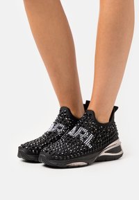 Schwarze Sportschuhe mit einer genoppten Textur, akzentuiert mit glänzenden Strasssteinen, die "DIVA" buchstabieren, und einer gepolsterten Sohle für Unterstützung.