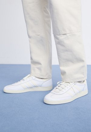 Personne portant des baskets blanches et un pantalon beige clair debout sur un sol recouvert d'un tapis bleu.