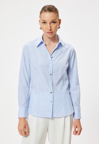 Camisa de botones a rayas en azul claro con cuello, mangas largas y botones en tono plateado. Tela suave y diseño ajustado, con un dobladillo recto.