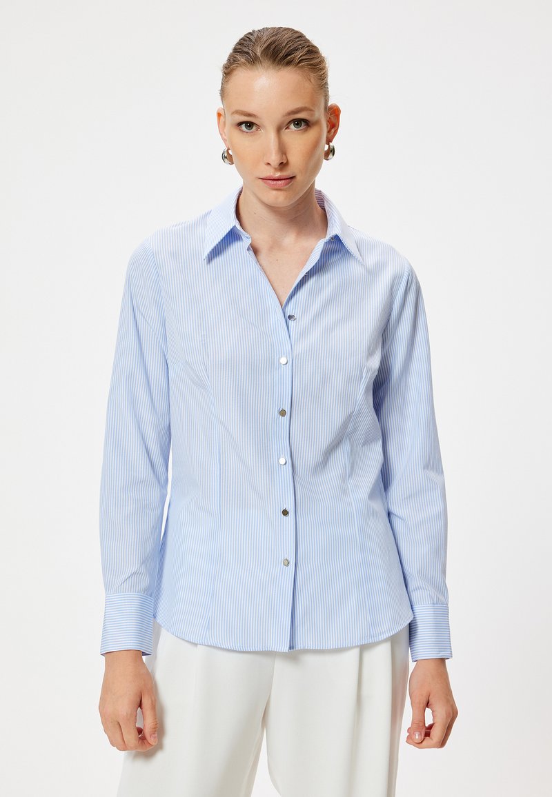 Camisa de botones a rayas en azul claro con cuello, mangas largas y botones en tono plateado. Tela suave y diseño ajustado, con un dobladillo recto.
