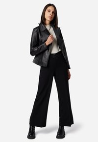 Schwarze Lederjacke mit gesteppter Schulterpartie, kombiniert mit hochgeschnittenen schwarzen weiten Hosen und schwarzen Stiefeletten. Neutrales Top darunter.