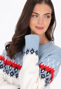 Stickad tröja med blå kropp, vita ärmar och mönstrade detaljer i röd och marinblå. Ribbad mudd och mjuk textur.