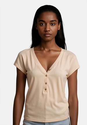 Donna con pelle medio-scura e lunghi capelli neri indossa una maglietta beige a maniche corte con scollo a V e tre bottoni, e jeans azzurri chiari.