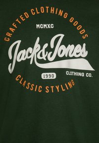 T-shirt in cotone verde con un grande logo bianco e arancione riportante il testo "Jack & Jones," fondato nel 1990, e un design classico.