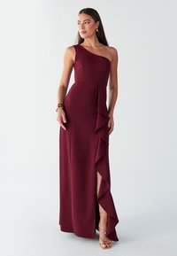 Burgundska večerna obleka z eno naramnico, z visokim razporkom na stegno in nabrano plastjo. Gladek material s prilegajočim se silhueto. Zlate sandale z naramnicami.