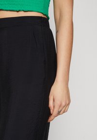 Pantalons larges noirs à texture exposés ; dotés d'une taille élastique et de poches latérales. Associés à un top court vert.