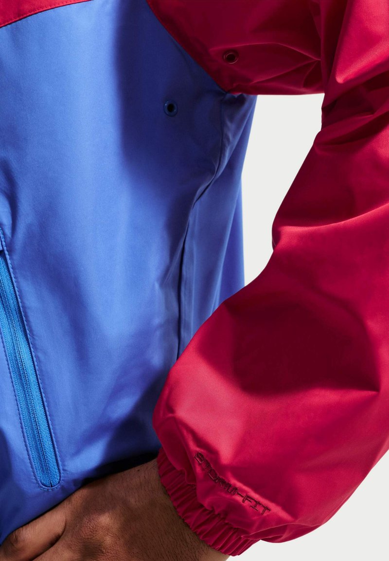 Rode en blauwe jaszoom met elastische manchet en ritszak op blauwe stof, gedragen door een persoon met zichtbare hand en pols.