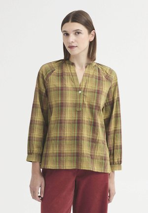 Femme aux cheveux bruns et raides portant un chemisier à carreaux jaunes et verts à manches longues avec un seul bouton et un pantalon rouge.