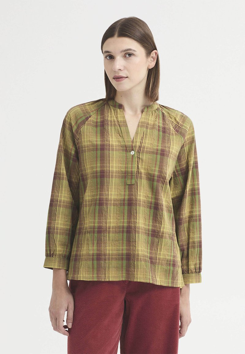 Femme aux cheveux bruns et raides portant un chemisier à carreaux jaunes et verts à manches longues avec un seul bouton et un pantalon rouge.