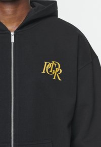 Felpa nera con zip realizzata in cotone morbido, con un design del logo dorato sulla parte sinistra del petto. Tessuto testurizzato e vestibilità standard.
