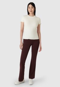 Bílý žebrovaný top s krátkým rukávem a scalloped okraji, spárovaný s bordovými rozšířenými kalhotami. Modelka má na sobě bílé tenisky, neutrální pozadí.