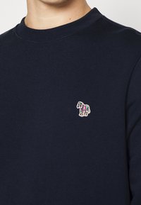 Persona che indossa una maglietta a girocollo di colore blu scuro con un piccolo logo ricamato a strisce multicolore di una zebra sul lato sinistro del petto.