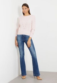 Suéter de punto trenzado de color rosa claro con cuello redondo y detalles de calado, combinado con jeans acampanados azules y zapatos de tacón desnudo.