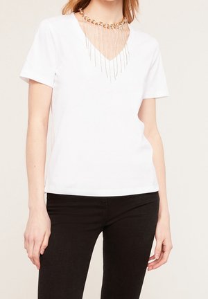 T-shirt blanc à manches courtes et col en V, featuring un collier en chaîne dorée avec des chaînes argentées en cascade, sur un fond neutre.