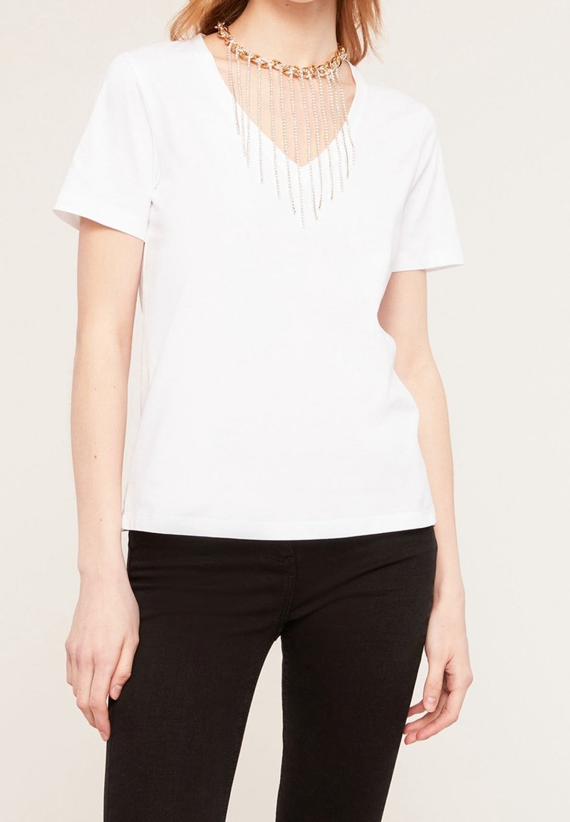 T-shirt bianco a maniche corte con scollatura a V, caratterizzato da una collana con catena d'oro e catene d'argento che cadono, posizionato su uno sfondo neutro.