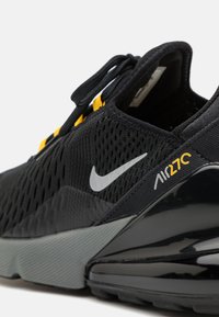 Svart träningsskor med meshuppbyggnad, gula accenter, en silverfärgad Nike swoosh och en blanka svarta luftkudde sula. Har en strukturerad design.