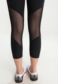 Black leggings with mesh panels, textured finish, and a mid-calf length, paired with white and black athletic shoes.
