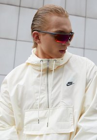 Hvid Nike vindjakke med hætte, frontlomme og snore. Model iført solbriller med tonede linser og et slankt design.