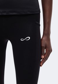 Gros plan sur une personne portant un legging de sport noir avec un logo symbole infini blanc sur la cuisse et une poche latérale, main reposant le long de la jambe.