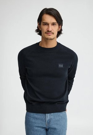 Uomo con capelli scuri di media lunghezza che indossa un maglione nero Polo Club e jeans blu, in piedi con le mani dietro la schiena su sfondo bianco.