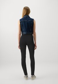 Jeans prsluk bez rukava s prilagođenim oblikom, tamnoplave boje i neuređenim rubovima u kombinaciji s crnim tajicama i svijetlimtenisicama.
