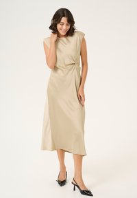 Abito in satin beige con maniche a raglan e dettaglio annodato in vita, abbinato a décolleté nere con punta affilata.