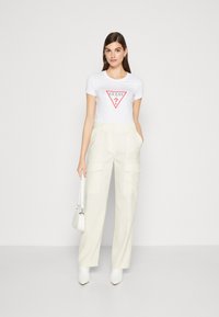 Hvid T-shirt med et rødt trekantet logo grafik, parret med brede cremfargede bukser med to forreste lommer. Modellen holder en lille hvid taske.